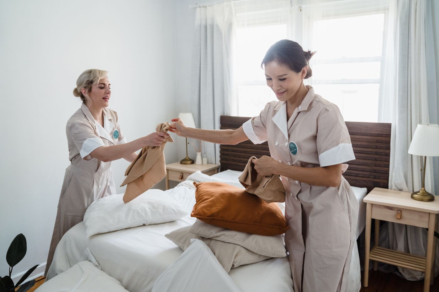 Two cleaners in uniform making a bed in a bright bedroom.
