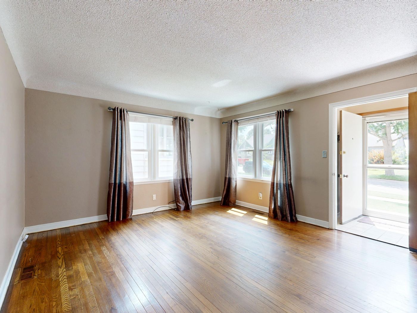 An empty living room with bare hardwood floors and dated curtains.
